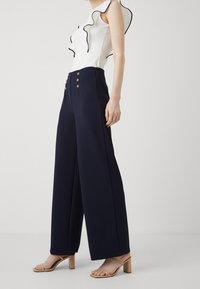 Navy wide-leg trousers in soft fabric, featuring a high waist with three gold button accents. Paired with a white top with black ruffles.