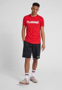 Rode t-shirt met "hummel" logo, zwarte shorts en grijze sneakers met rode accenten. Witte en zwarte gestreepte sokken maken de outfit compleet.