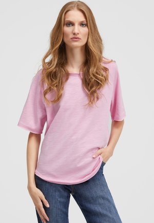 Frau mit langen, welligen, hellbraunen Haaren trägt ein lockeres, rosa Kurzarmhemd und blaue Jeans, steht mit einer Hand in der Tasche vor einem weißen Hintergrund.