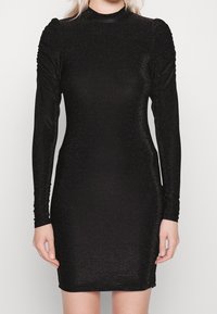 Svart långärmad klänning i glittrande material, med en figurnära silhuett och rynkade detaljer längs axlarna och ärmarna.