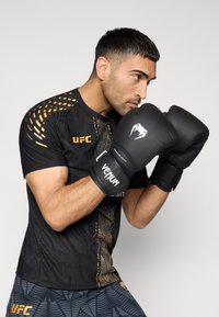 Μαύρο κοντομάνικο πουκάμισο με πορτοκαλί λεπτομέρειες και υφή, μαύρα γάντια πυγμαχίας με λευκό λογότυπο. Μοντέλο σε στάση πυγμαχίας.