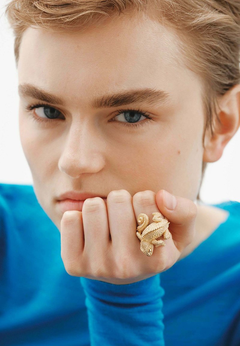 Anillo dorado con forma de lagarto, que presenta escamas texturizadas y detalles en espiral, llevado en la mano que reposa sobre una manga azul.