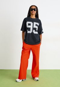 Svart oversized t-skjorte med hvit "95" grafikk, kombinert med knallorange vide bukser. Hvit og svart sneakers, accentuert med solbriller.