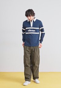 Marineblauw rugbyshirt met lange mouwen, een witte kraag en horizontale crèmekleurige strepen; voorzien van het "ellesse" logo. Gecombineerd met olijfgroene cargobroeken en witte sneakers.