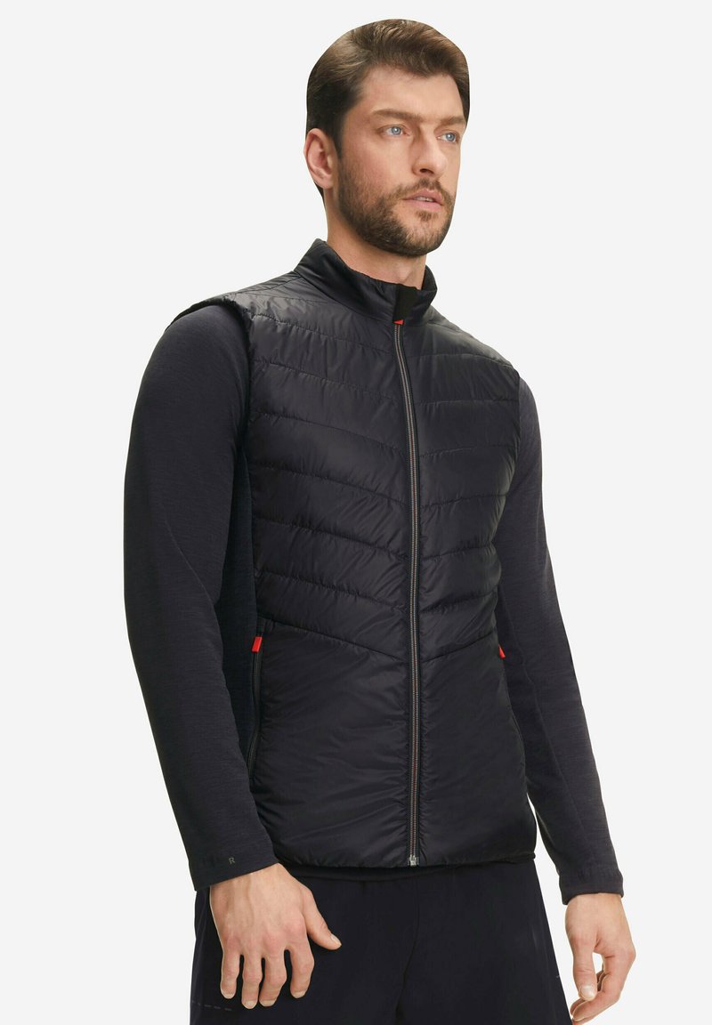 Uomo che indossa un giubbotto senza maniche trapuntato nero sopra una maglia nera a maniche lunghe, in piedi davanti a uno sfondo chiaro e semplice.