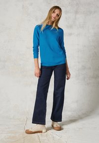 Maglione blu a maglia con maniche lunghe, abbinato a jeans neri a gamba larga e sneakers beige con un accento testurizzato. Sfondo neutro.