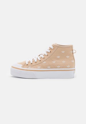 Beżowy wysokiego kroju sneaker z białymi sznurówkami, gumową podeszwą i białymi haftowanymi logo z koniczynką na materiałowej cholewce, widok z boku na białym tle.