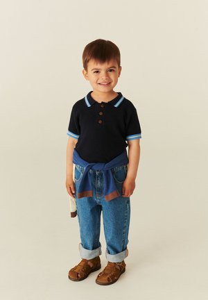Jeune garçon souriant, portant un polo noir, un jean bleu avec des revers, un pull bleu noué autour de la taille et des sandales marron, debout sur un fond uni.