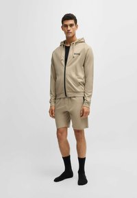 Sudadera cremosa con cremallera, logo negro y bolsillos, combinada con pantalones cortos a juego. El modelo lleva calcetines negros. Tejido suave, diseño sencillo.