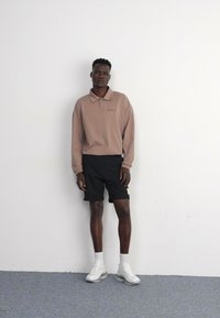 Camisa polo de manga larga marrón con cuello, pantalones cortos negros y zapatillas blancas. Diseño minimalista, colores sólidos y corte relajado.
