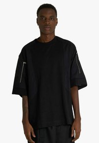 T-shirt nera oversize con design a pannelli, maniche corte e tasche con zip su entrambe le braccia. Realizzata in tessuto liscio con finitura opaca.