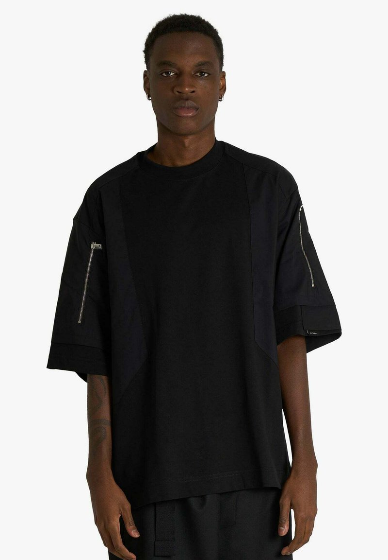 T-shirt nera oversize con design a pannelli, maniche corte e tasche con zip su entrambe le braccia. Realizzata in tessuto liscio con finitura opaca.