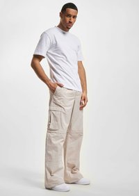 T-shirt blanc à manches courtes associé à un pantalon cargo beige clair avec plusieurs poches. Le mannequin complète la tenue avec des baskets blanches.
