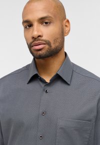 Grauhemd mit strukturiertem Muster, ausgestattet mit einer Tasche auf der linken Seite, spitzen Kragen und dunklen Knöpfen mit roten Akzenten.