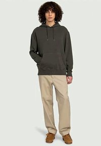Jovem com cabelo encaracolado, vestindo um hoodie verde escuro, calças bege e sapatos castanhos, de pé com uma das mãos no bolso contra um fundo simples.