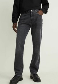 Persona con botas negras, jeans rectos desgastados de color negro, sudadera negra, con la mano derecha en el bolsillo, contra un fondo liso.
