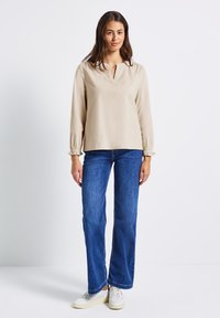 Frau steht, trägt eine beigefarbene Bluse mit langen Ärmeln, blaue Jeans und weiße Sneaker vor einem schlichten weißen Hintergrund.