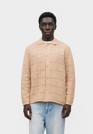 Muž oblečený v béžovom pletenom kardigane na gombíky so štvorcovým vzorom a svetlo modrých džínsoch, stojaci pred jednoduchým svetlým pozadím.
