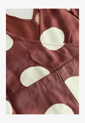 Kleid aus maronenfarbener Stoff mit großen, cremefarbenen Punkten. V-Ausschnitt mit strukturierter Optik und überlappenden Stoffabschnitten.