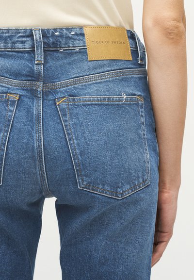 Κοντινό πλάνο ατόμου που φοράει μπλε τζιν με πίσω τσέπες και δερμάτινη ετικέτα της μάρκας "Tiger of Sweden" στη μέση.