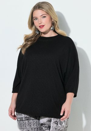 Femme aux longs cheveux blonds portant un haut noir à manches trois-quarts, un pantalon gris et blanc à motifs, et de grandes boucles d'oreilles argentées, sur un fond uni.