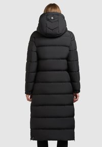 Svart pufferjacka med lång design, med en kviltad textur, huva och en knappdetalj på baksidan.