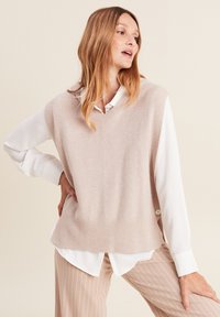 Lichtroze gebreide vest met een V-hals en geribbelde zoom, gelaagd over een wit shirt, in combinatie met beige gestreepte broek.