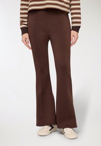 Pantaloni marroni svasati in tessuto morbido, con dettagli a cucitura e una texture liscia. Indossati con sneakers di colore chiaro. Simpatica maglia a righe visibile.