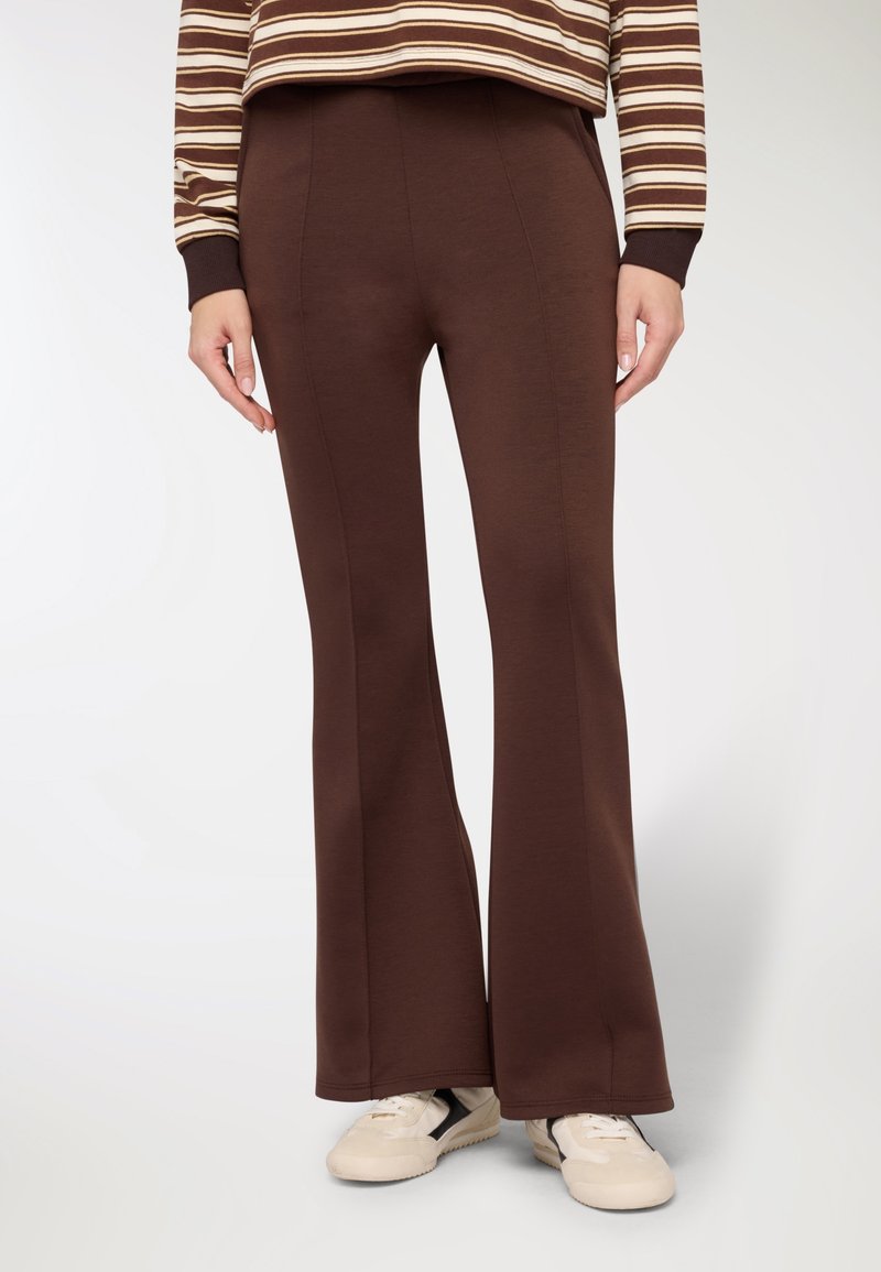 Pantaloni marroni svasati in tessuto morbido, con dettagli a cucitura e una texture liscia. Indossati con sneakers di colore chiaro. Simpatica maglia a righe visibile.