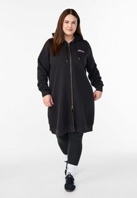 Sort quiltet hætteklæde med lynlås, hætte, guldfarvet lynlås og ribstrikkede manchetter. Modellen bærer den med sorte leggings og sorte sneakers.