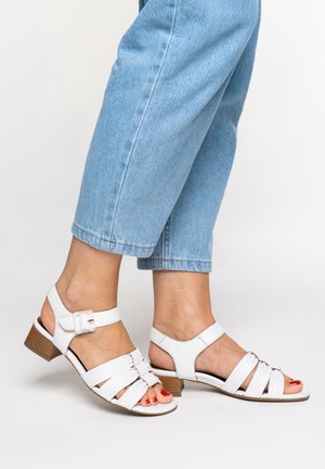 Feet wearing white leather heeled sandals with buckles and open toes, paired with light blue cropped denim jeans.