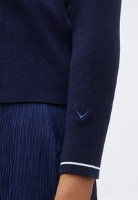 Μανίκι πουλόβερ σε σκούρο μπλε με ριμπ ύφασμα, διακοσμημένο με μικρό κεντητό σχήμα σε σχήμα διπλού βέλους και λεπτομέρεια λευκής ρίγας, συνδυασμένο με υφασμάτινη φούστα σε σκούρο μπλε χρώμα.