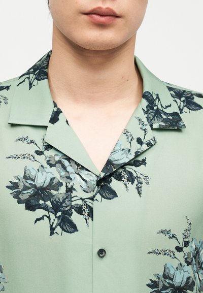 Άνδρας που φοράει ανοιχτό πράσινο πουκάμισο με μαύρο λουλουδάτο σχέδιο και ανοιχτό γιακά, εμφανίζοντας μόνο τον λαιμό και το κάτω μέρος του προσώπου.