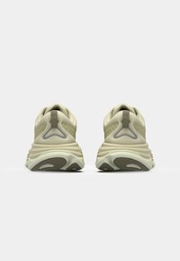Beige treningssko med en teksturert overdel, buet hæl og fleksibel såle. Detaljene inkluderer en grå logo og en liten løfletapp bak.