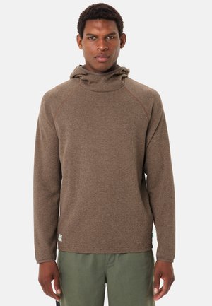 Uomo di fronte che indossa un pullover con cappuccio marrone a maniche lunghe e pantaloni verde oliva su uno sfondo chiaro e uniforme.