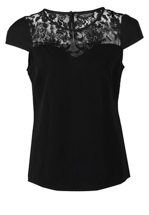 Kobieta w czarnej koronkowej bluzce, niebieskich obcisłych dżinsach, czarnych spiczastych balerinach, niosąca czarną torebkę na ramię z łańcuszkowym paskiem.