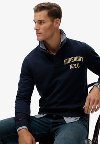 Suéter azul marino con un cuello alto, que presenta el texto crema "SUPERDRY N.Y.C." Llevado sobre una camisa de cuadros en azul claro. Jeans de mezclilla azul.