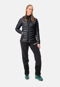 Schwarze isolierte Jacke mit gestepptem Design, durchgehendem Reißverschluss und zwei Taschen, kombiniert mit schwarzen Hosen und grünen Stiefeln. Der Hintergrund ist rein weiß.