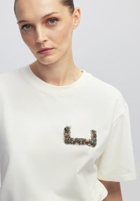 Witte katoenen t-shirt met een textuur, met een juweelversiering in een geometrisch patroon op de borst, met korte mouwen en een ronde hals.