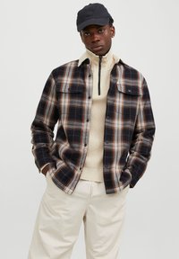 Geruit shirt in donkerblauw, bruin en beige; twee borstzakken; gecombineerd met een crème trui met hoge hals. Model draagt een zwarte pet.