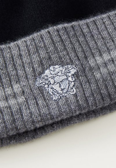Berretto grigio in maglia con una texture a coste. Presenta un'emblema ricamato di colore azzurro chiaro. Il design include un bordo piegato.