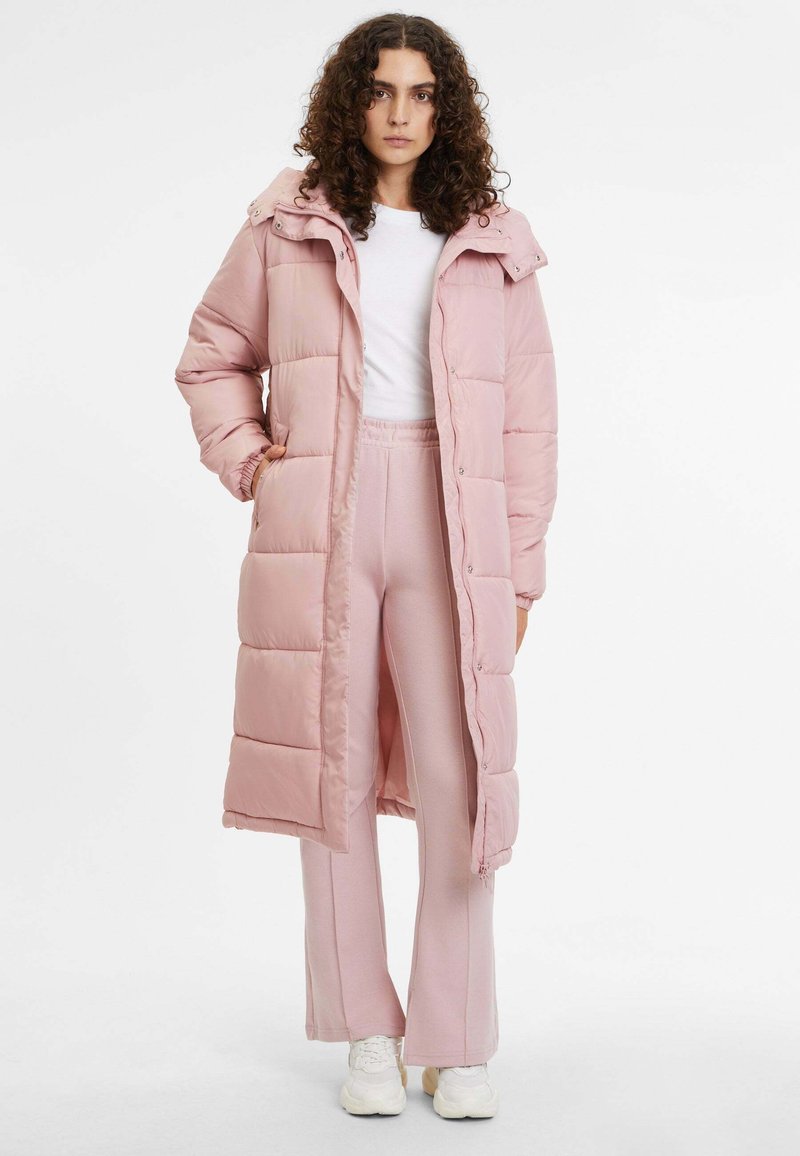 Lange roze pufferjas met een capuchon, voorzien van gequiltte delen en drukknoopsluitingen, gecombineerd met bijpassende roze joggingsbroek en witte sneakers.