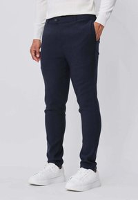 Pantalon bleu marine à coupe slim en tissu doux, doté de poches latérales et d'une fermeture à bouton unique. Porté avec des baskets blanches pour le contraste.