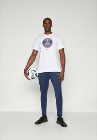 Hvid Nike t-shirt med et rundt Paris-logo, kombineret med marineblå træningsbukser og sorte fodboldsko. Holder en mønstret fodbold.
