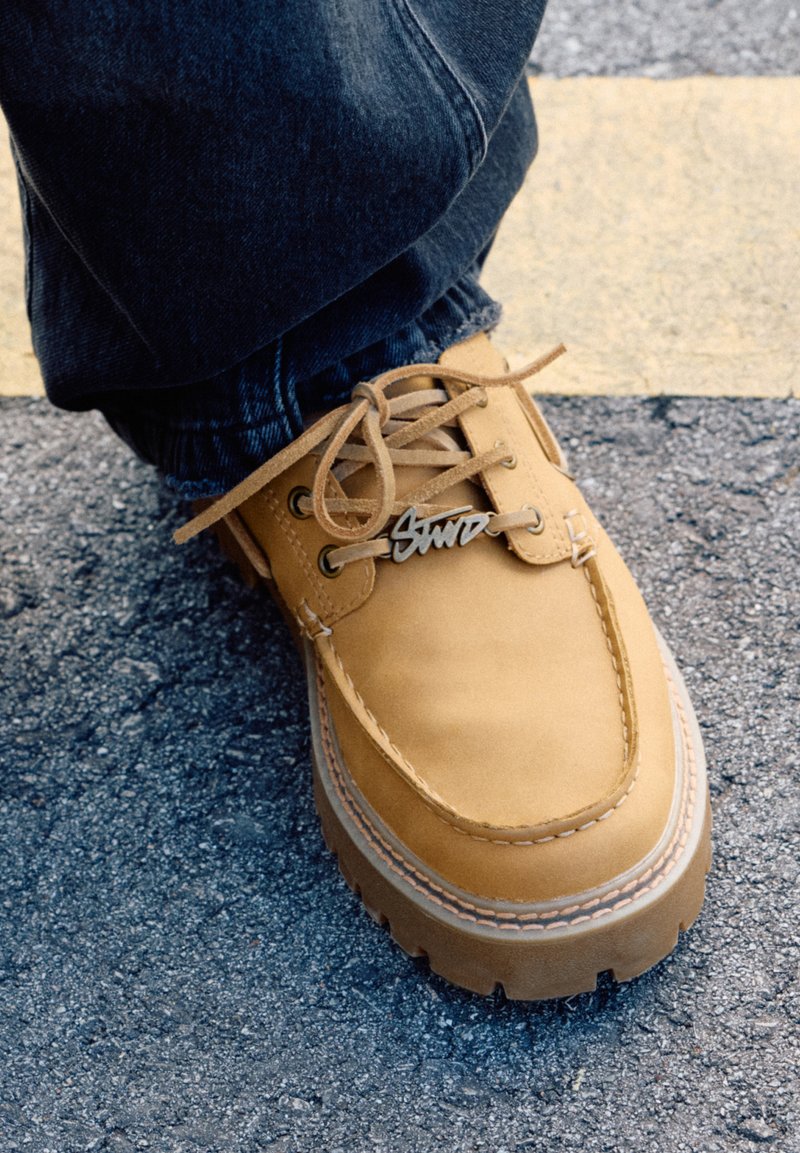 Scarpa da barca in pelle marrone chiaro con suola robusta, cuciture bianche a contrasto e hardware incisa "Gnu" sui lacci. Texture e design solidi.