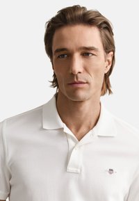 Polo blanc en tissu texturé, avec une patte de boutonnage à deux boutons et un petit logo brodé sur la poitrine gauche. Design décontracté et ajusté.
