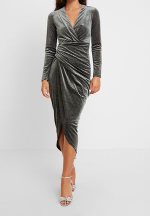 Šedé sametové šaty s dlouhými rukávy, výstřihem do V, nařasenými detaily a rozparkem nad stehno, doplněné o stříbrné sandály na podpatku.