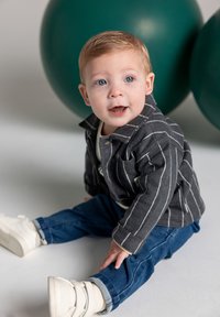 Toddler with light hair wearing a striped jacket, jeans, and white sneakers, sitting on the floor with large green balloons behind.