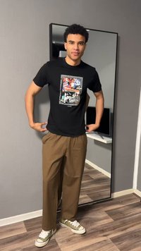 Νέος άνδρας με μαύρο T-shirt με σχεδιασμό, καφέ παντελόνια και πράσινα αθλητικά παπούτσια, στέκεται μπροστά σε έναν καθρέφτη ολόσωμο σε ένα δωμάτιο.