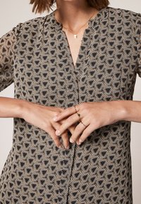 Chemise noire et dorée à motif géométrique, col en V et manches courtes. Les mains sont croisées devant, mettant en valeur des bagues.